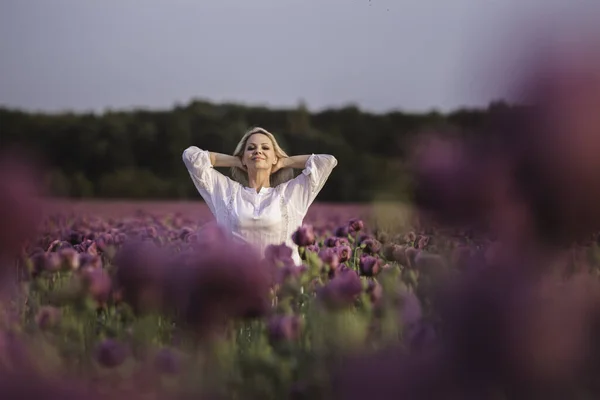 Schöne, glückliche Frau mit langen Haaren im weißen Kleid, die einsam im Flieder-Mohnblumen-Feld spaziert — Stockfoto