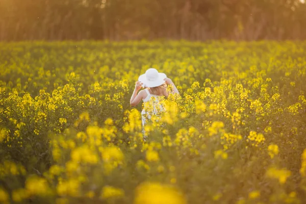 Imádnivaló kislány fehér ruhában és kalapban fut a tavaszi mezőn sárga virágok, nemi erőszak — Stock Fotó