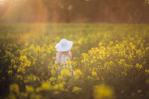 Uma menina borrada de vestido branco a correr pelo campo de violação ao pôr-do-sol. Brilho intencional do sol, foco da lente em flores de estupro — Fotografia de Stock