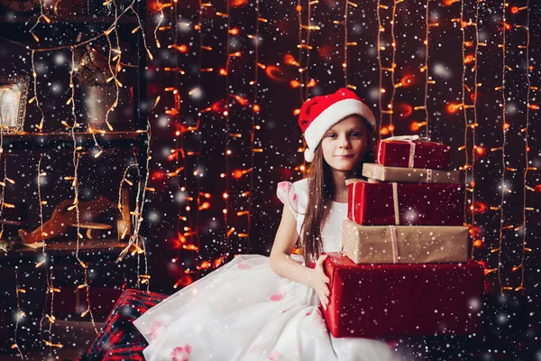 Härlig flicka i Santa Claus hatt och klänning håller gäng julklappar under snöflingor. — Stockfoto