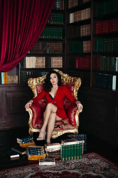 Retrato de morena em vestido vermelho sentado em poltrona no interior da biblioteca . — Fotografia de Stock