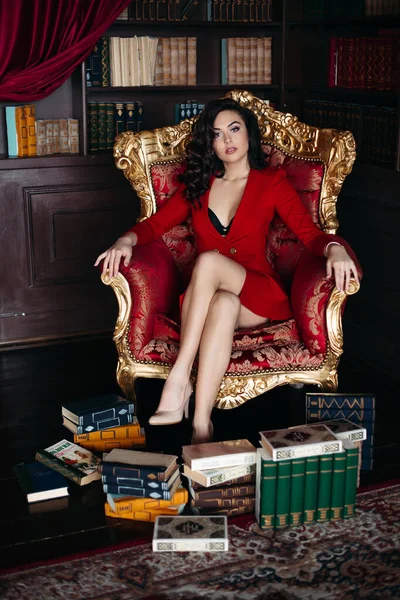 Portret brunetki w czerwonej sukience siedzącej na fotelu w bibliotece wnętrza. — Zdjęcie stockowe