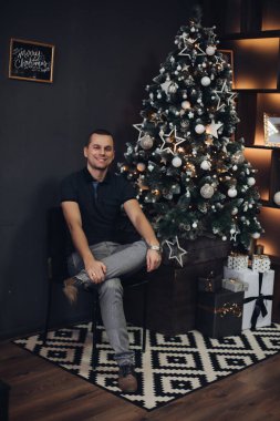 Aydınlanmış Noel ağacına karşı yakışıklı bir adam. Elleri çenesinde ve düşünceli bakışlarıyla başka tarafa bakan şık bir saat..