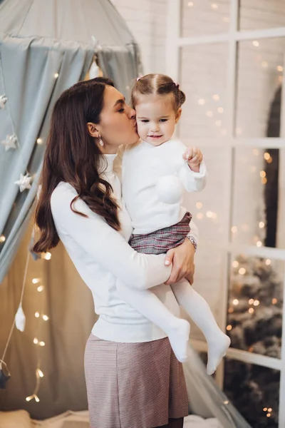 Mother kissing her daughter in cheek. room decorated for Christmas.