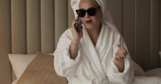 Mujer de moda sonriendo hablando con el teléfono inteligente después de la ducha acostado en la cama de tiro completo — Vídeos de Stock