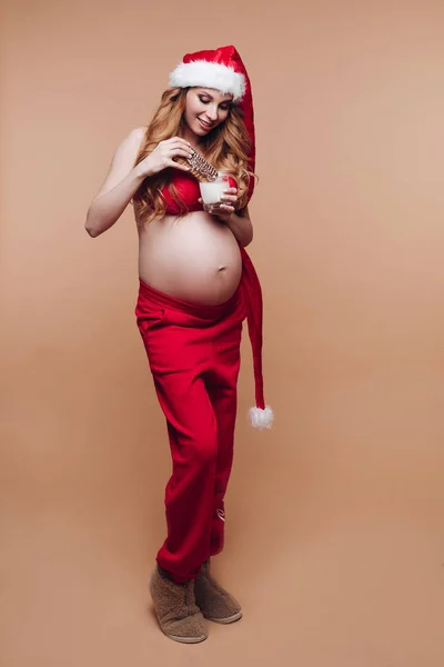 Lachend zwanger meisje in Santa Claus pak eten peperkoek koekjes met melk hebben plezier — Stockfoto