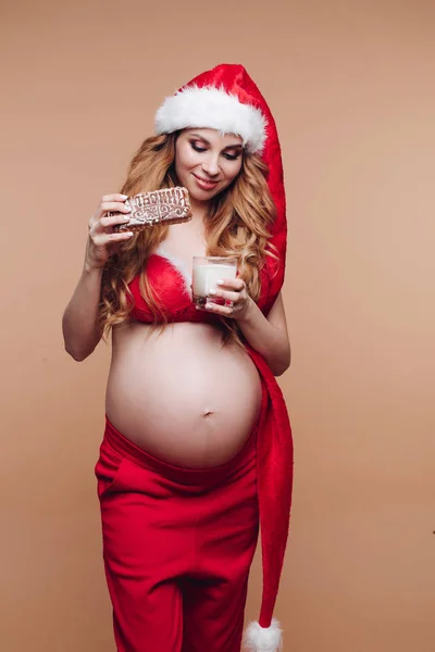 Sonriendo chica embarazada en traje de Santa Claus comer galletas de jengibre con leche divertirse — Foto de Stock
