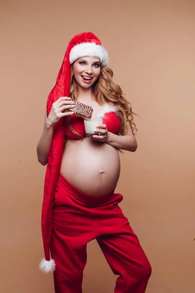 Улыбающаяся беременная девушка в костюме Санта-Клауса ест пряничное печенье с молоком, веселясь — стоковое фото
