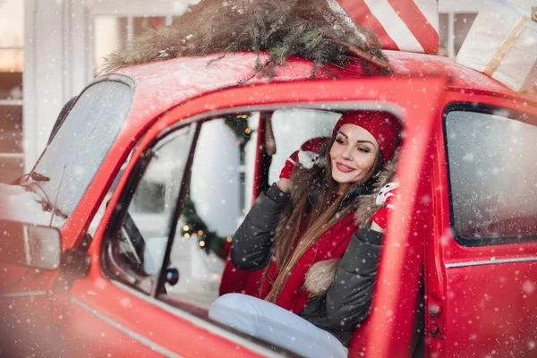 눈송이로 둘러싸인 레드 빈티지 차에서 포즈를 취하며 멋진 겨울 소녀가 웃는 모습 — 스톡 사진