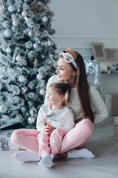 Feliz madre festiva e hija abrazándose sentados juntos en la decoración de hermoso árbol de Navidad —  Fotos de Stock