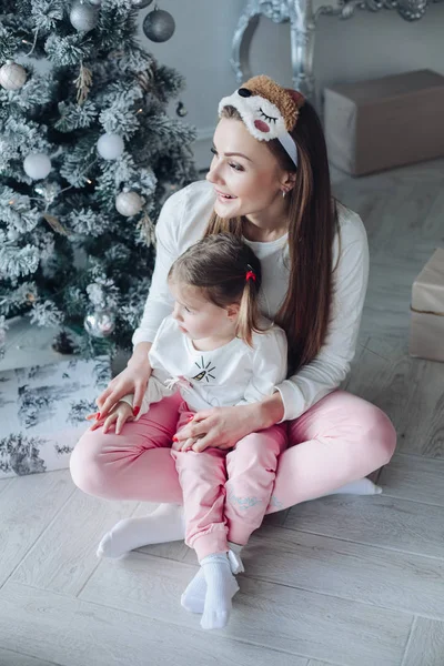 Feliz madre festiva e hija abrazándose sentados juntos en la decoración de hermoso árbol de Navidad —  Fotos de Stock
