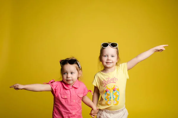 两个快乐而漂亮的小女孩戴着太阳镜，摆出孤零零的黄色画室背景 — 图库照片