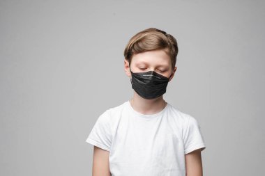 Genç bir çocuğun portresi gri arka planda siyah bir tıbbi maske takıyor.