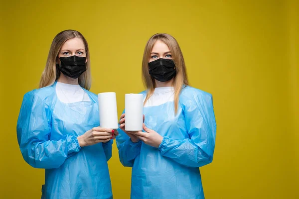 Молоді красиві дівчата в одноразових медичних сукнях і з масками на обличчях тримають мокрі антибактеріальні серветки, портрет ізольований на жовтому фоні — стокове фото
