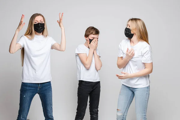 Portret van twee knappe meisjes met lang blond haar en een jonge jongen met zwarte medische maskers op hun gezicht paniek geïsoleerd op grijze achtergrond — Stockfoto