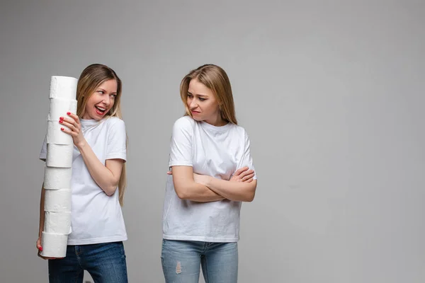 Portrait de deux belles filles aux longs cheveux blonds, l'une d'elles tient beaucoup de papier toilette et l'autre est offensée isolée sur fond gris — Photo