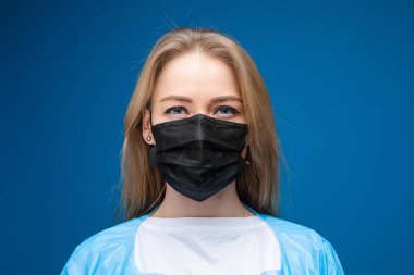 Yüzünde tıbbi maske olan uzun sarı saçlı güzel bir kızın portresi, mavi arka planda izole edilmiş bir resim.