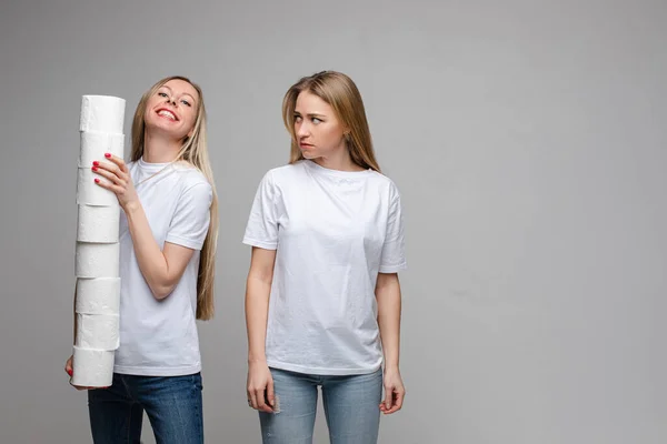 Portret van twee knappe meisjes met lang blond haar, een van hen heeft veel toiletpapier en de ander is beledigd geïsoleerd op grijze achtergrond — Stockfoto