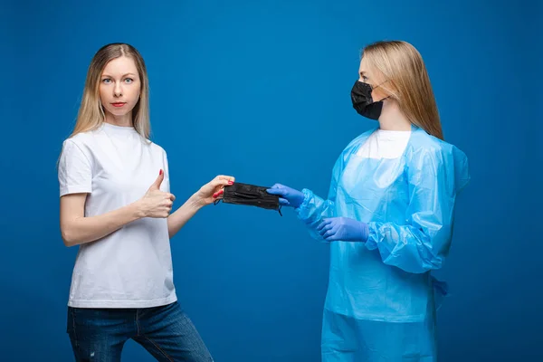 Unga vackra flickor i en engångs medicinska klänningar och med masker, porträtt isolerad på blå bakgrund — Stockfoto