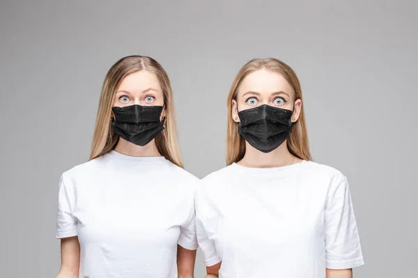 Portret van twee knappe meisjes met lang blond haar met zwarte medische maskers op hun gezicht geïsoleerd op grijze achtergrond — Stockfoto