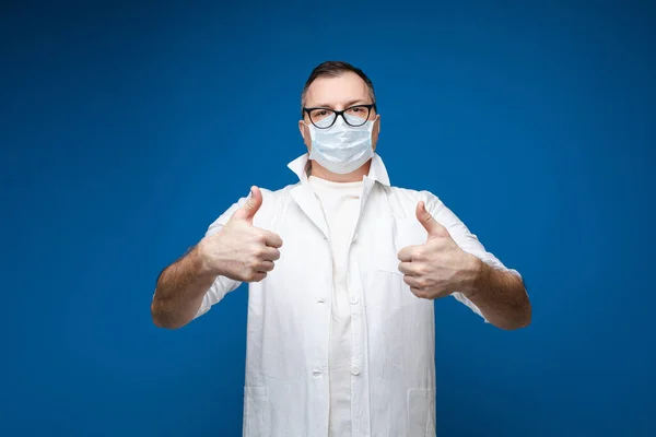Сильний чоловік в окулярах, біла медична сукня і маска стоїть і показує, як, картина ізольований синій фон — стокове фото