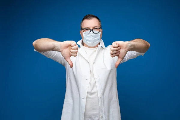 Сильний чоловік в окулярах, біла медична сукня і маска стоїть і показує відмінність, картина ізольований синій фон — стокове фото
