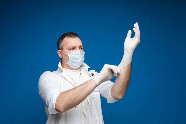 Сильний чоловік в окулярах, біла медична сукня і маска стоїть і носить рукавички, картина ізольована на синьому фоні — стокове фото