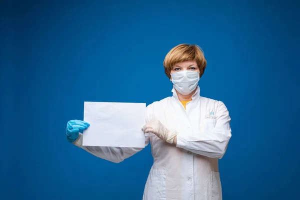 Vacker kaukasiska kvinna med medicinsk mask håller ett vitt papper, porträtt isolerad på blå bakgrund — Stockfoto