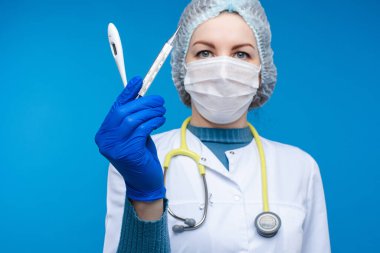 Beyaz elbiseli, maskeli, fonendoskoplu ve tıbbi şapkalı güzel kadın doktor elektrikli termometreleri gösteriyor. Mavi arka planda izole edilmiş bir resim.