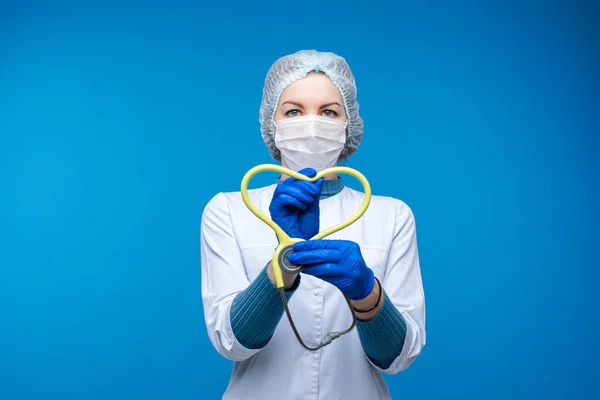 Красива жінка-лікар в білому медичному одязі, маска тримає фонендоскоп і показує серце, зображення ізольоване на синьому фоні — стокове фото
