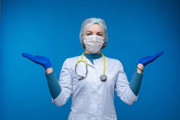 Vacker kvinna läkare i vita medicinska kläder, mask, fonoskop och medicinsk hatt vet inte vad man ska göra, bild isolerad på blå bakgrund — Stockfoto