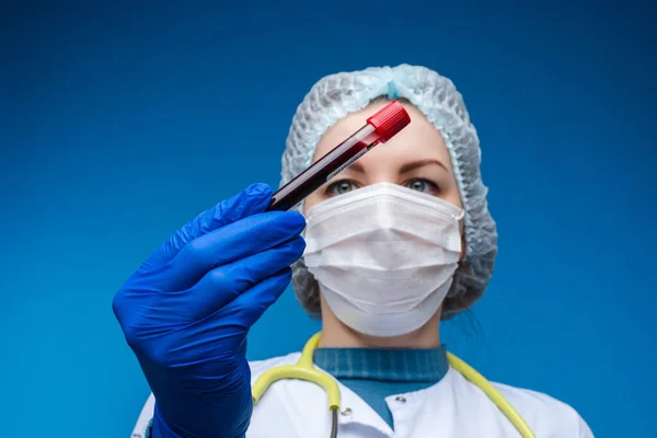 Красива жінка-лікар у білому медичному одязі, масці, фонендоскопі та медичному капелюсі показує кров у вітро, зображення ізольоване на синьому фоні — стокове фото