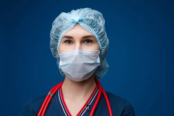 Красива жінка-лікар у білому медичному одязі, масці, фонендоскопі та медичному капелюсі, картина ізольована на синьому фоні — стокове фото