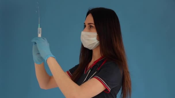 Beautiful woman doctor in white medical clothes, mask, blue gloves and phonendoscope on her shoulders isolated on blue background — Stock Video