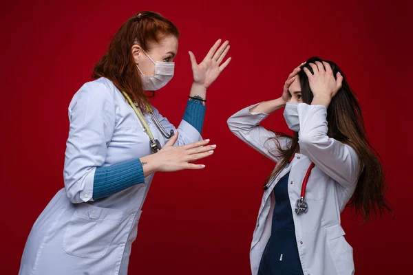 Porträtt av två kvinnliga läkare i medicinska kläder med fonendoskop runt halsen argumenterar, bild isolerad på röd bakgrund — Stockfoto