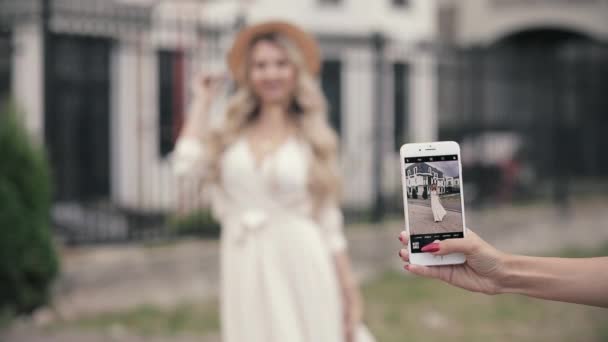 Mujer alegre con hermosos ojos azules grandes, pelo largo y claro, sombrero blanco en vestido blanco largo posa para la cámara del teléfono y sonríe — Vídeos de Stock