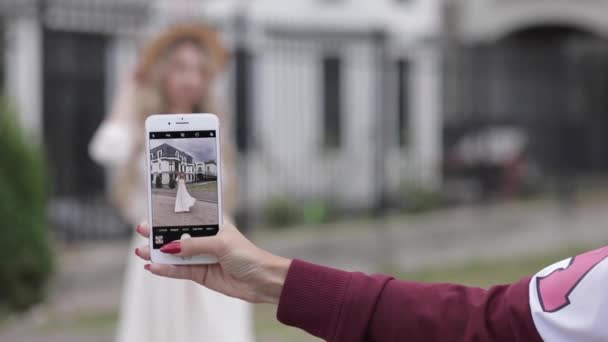 Femme gaie avec de beaux grands yeux bleus, longs cheveux blonds, chapeau blanc en robe blanche longue pose pour la caméra du téléphone et sourit — Video