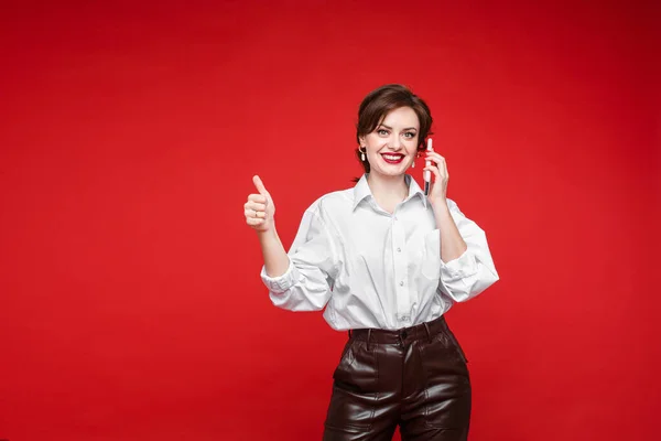 Bonita jovem conversa com sua amiga com um telefone celular e sorrisos, foto isolada no fundo vermelho — Fotografia de Stock