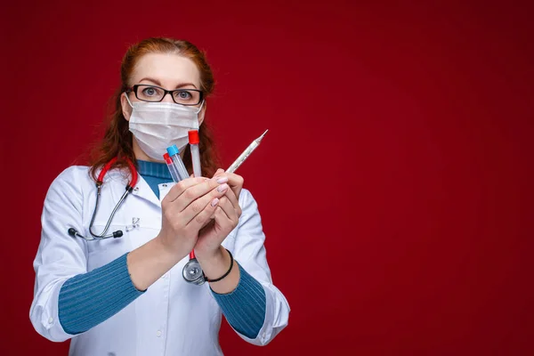 Портрет лікаря-жінки в медичному одязі з фонендоскопом навколо шиї тримає кілька пробірки і термометр, зображення ізольоване на червоному тлі — стокове фото