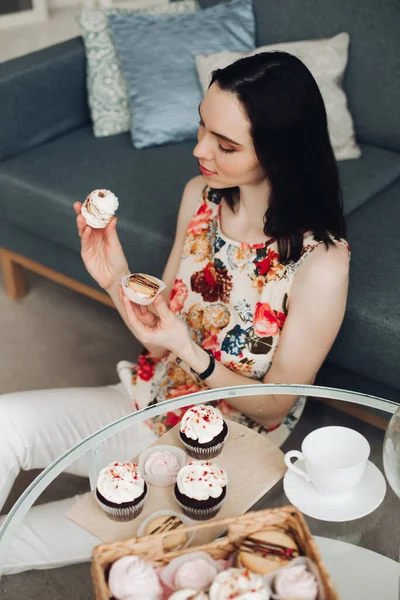 Wanita cantik dengan marshmallow di tangan. Dia duduk di meja kopi dengan cupcakes buatan sendiri. — Stok Foto