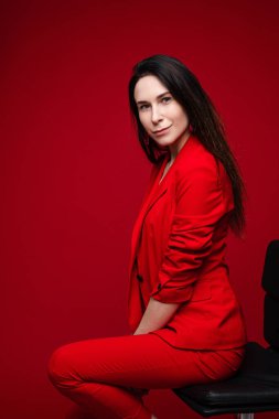 Beyaz, beyaz, uzun siyah saçlı, kırmızı takım elbiseli, siyah ayakkabılı, siyah sandalyede oturan ve kameraya poz veren bir kadın.