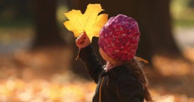 Siyah ceketli küçük güzel kız sonbahar parkındaki sarı yapraklara bakar ve sevinir.