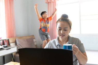 Evden çalışıyorum. Asyalı anne dizüstü bilgisayar kullanıyor, kahve içiyor ve oğlu gürültü yapıyor, sabahın köründe pencereden gelen güneş ışığıyla yatağa atlıyor..