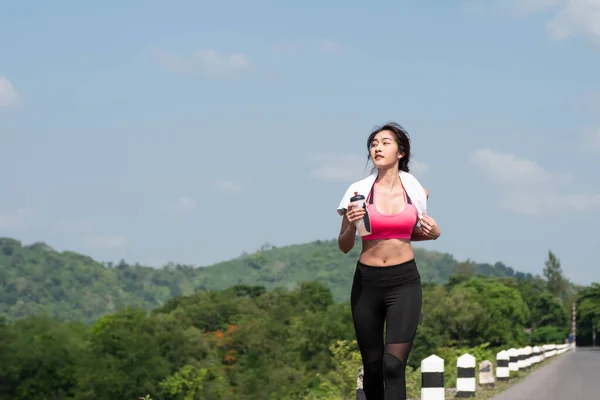 Ön manzara genç Asyalı kadın sabah güneşinde yolda koşuyor. Kadın koşucu doğada şişelenmiş su tutarak egzersiz yapıyor. Fitness ve sağlıklı yaşam tarzı