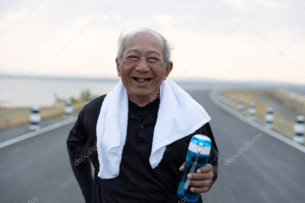 Front view Asian elderly exercise happy smile, Positive Emotion. Close up Senior man  holding water bottle on road in the morning sunshine. health lifestyle and exercise Concept.