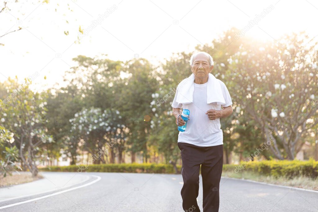 Front view Asian elderly exercise. Senior man holding water bottle running on road in public park that has sunshine in the evening. health lifestyle and exercise Concept.