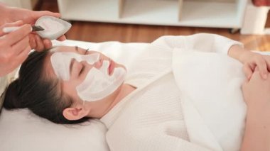 Kapalı kapılar ardında genç Asyalı bir kadın güzellik salonunda yüzüne fırça maskesi sürerek krem sürüyor. Cilt bakımı ve vücut bakımı. Dolly kaydırıcı