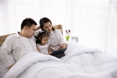 Mutlu Asyalı aile teknolojileri eğlence için kullanıyor. Baba, anne ve kız evdeki yataktaki dijital tablete bakıyorlar.