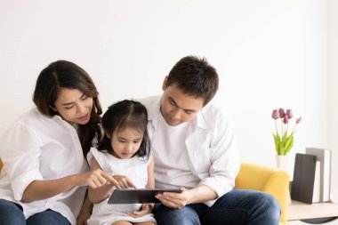 Mutlu Asyalı aile teknolojileri eğlence için kullanıyor. Baba, anne ve kız evdeki kanepede dijital tablete bakıyor. İletişim teknolojisi ve yaşam tarzı