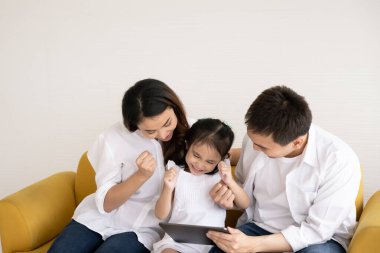 Mutlu Asyalı aile teknolojileri eğlence için kullanıyor. Baba, anne ve kız evdeki kanepede dijital tablete bakıyor. İletişim teknolojisi ve yaşam tarzı